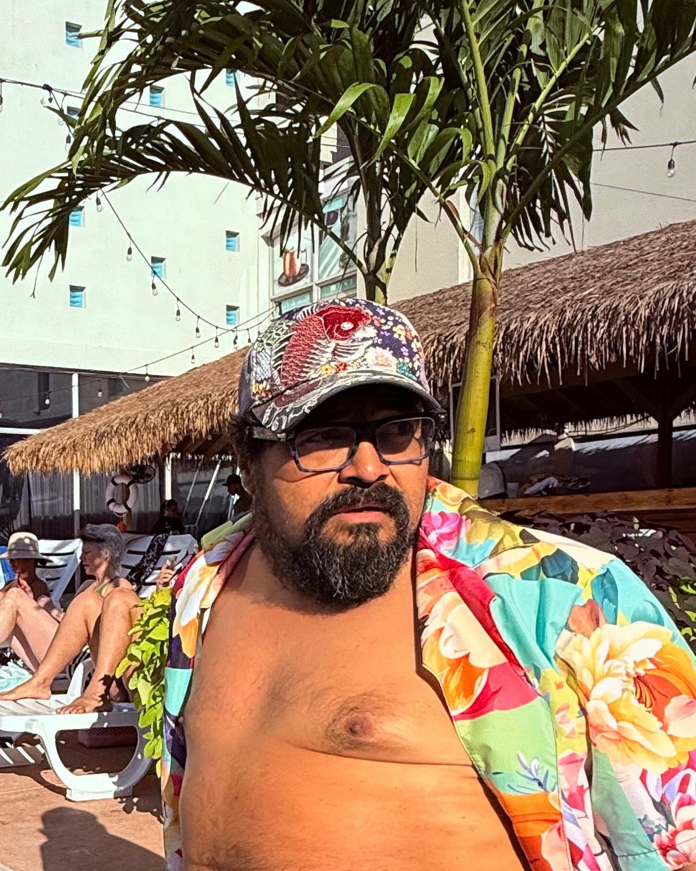 Man in colorful shirt and rainbow swim trunks sitting by a pool with palm trees and buildings in the background.