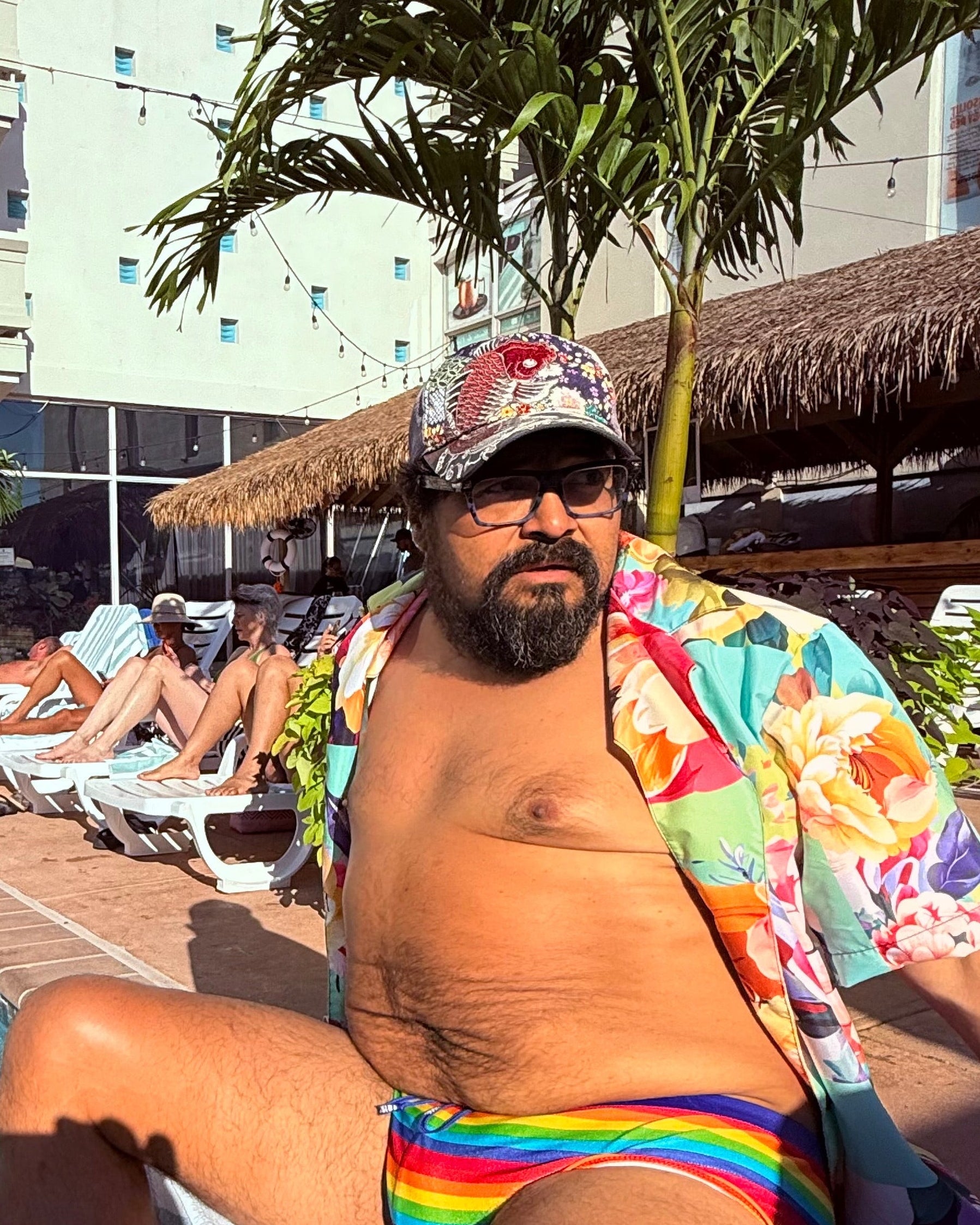 Man in colorful shirt and rainbow swim trunks sitting by a pool with palm trees and buildings in the background.