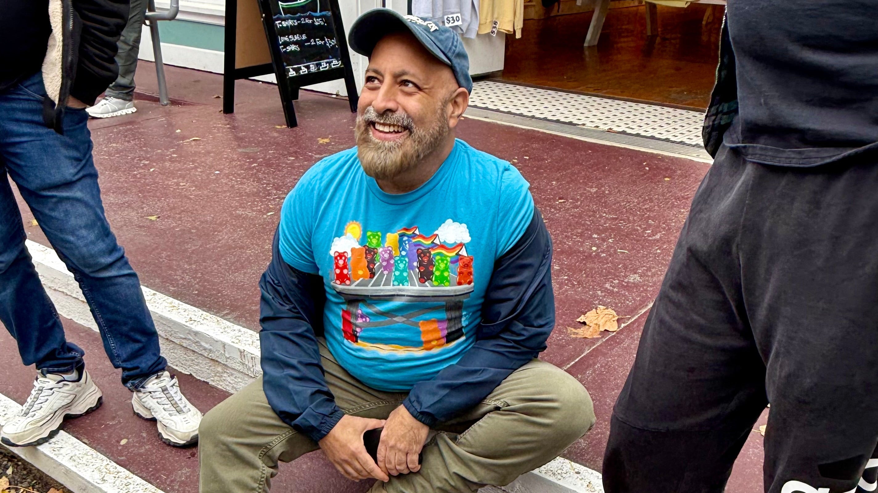 Man sitting on steps wearing a colorful graphic t-shirt with a store and people in the background.
