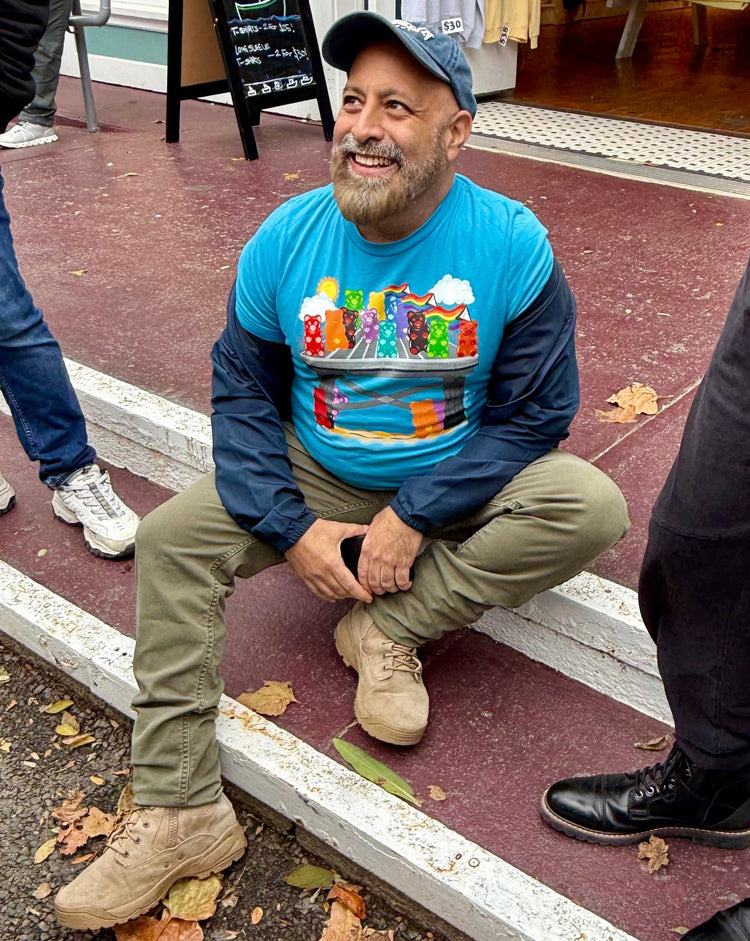 Man sitting on steps wearing a colorful graphic t-shirt with a store and people in the background.