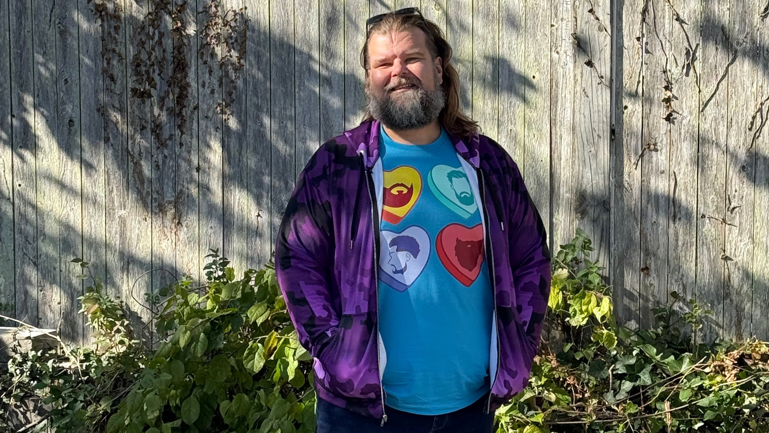 Person wearing a colorful shirt and purple jacket standing on a wooden deck with greenery in the background