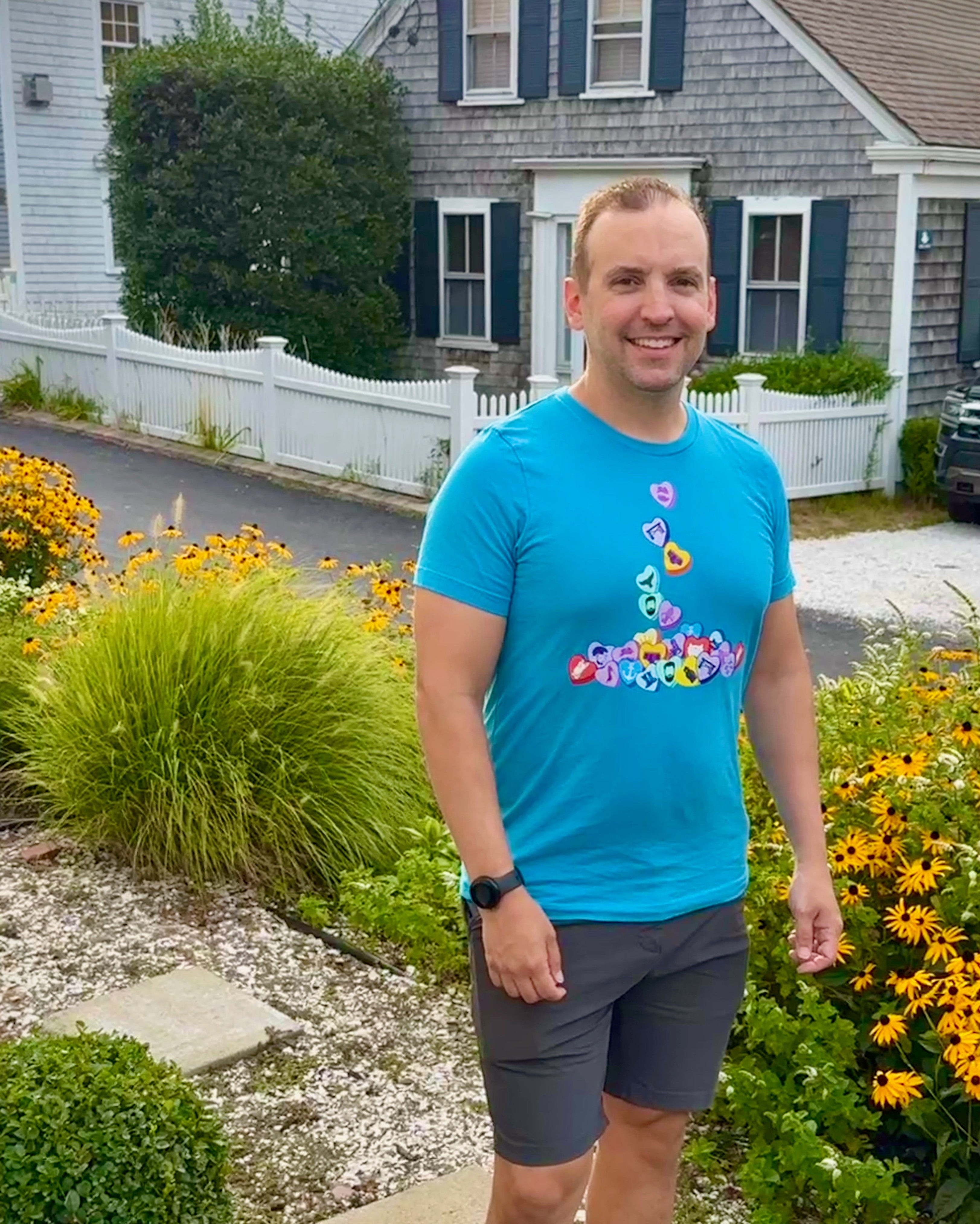 Man in a blue t-shirt with colorful design standing in front of a house with flowers and plants.