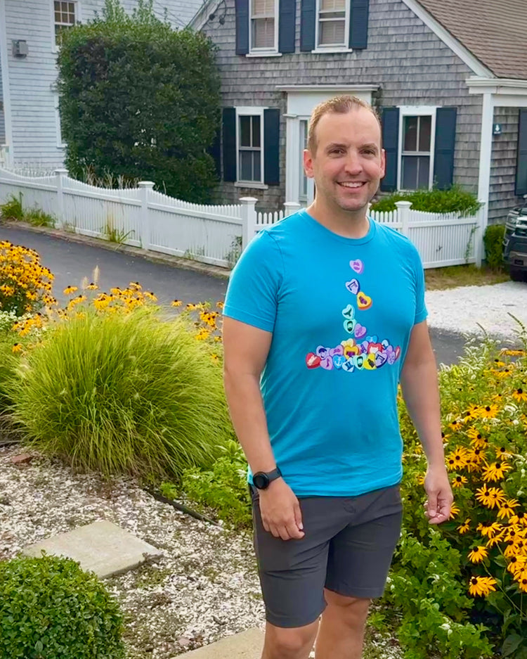 Man in a blue t-shirt with colorful design standing in front of a house with flowers and plants.