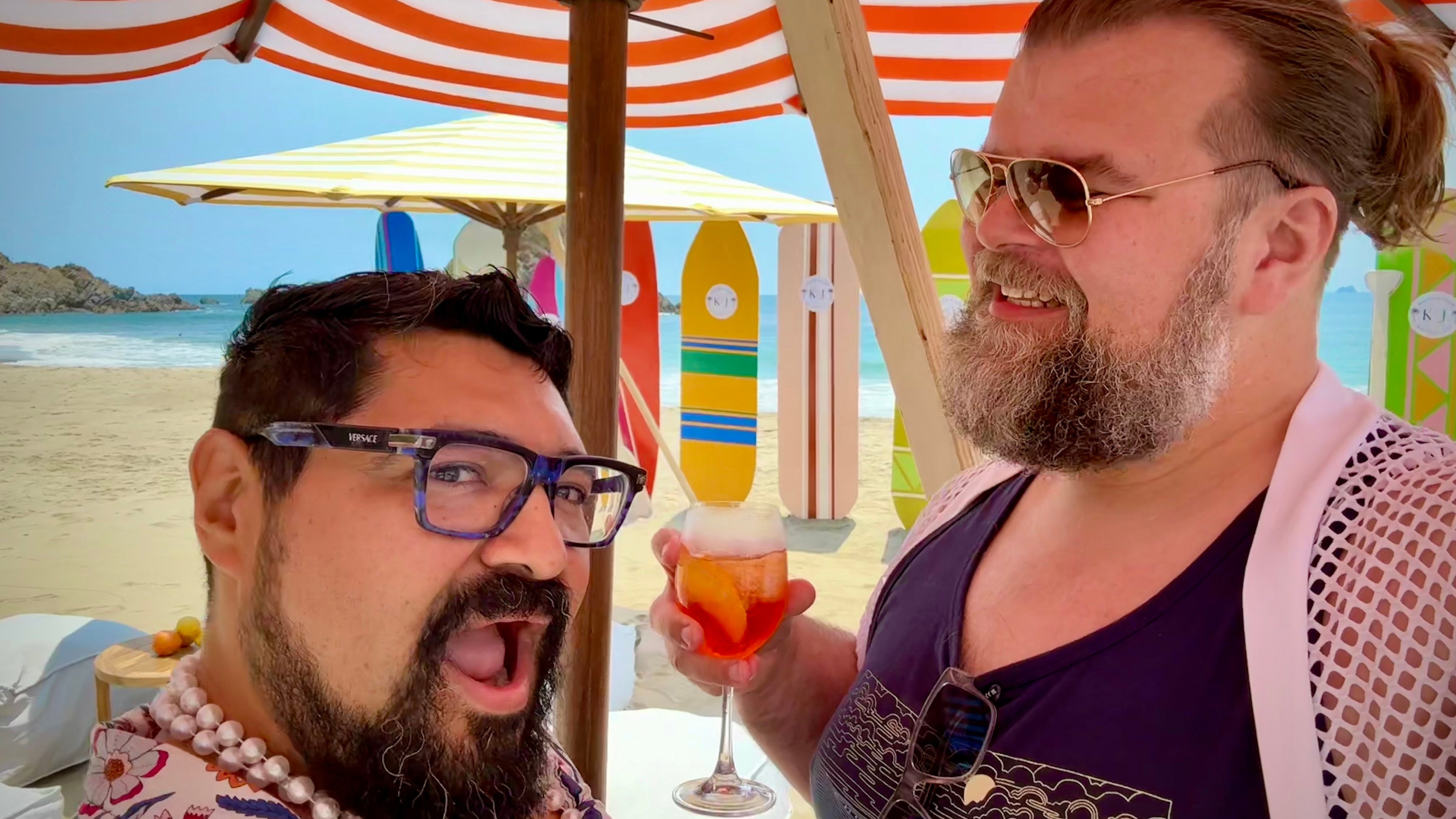 Two men on a beach with colorful umbrellas and a surfboard in the background.